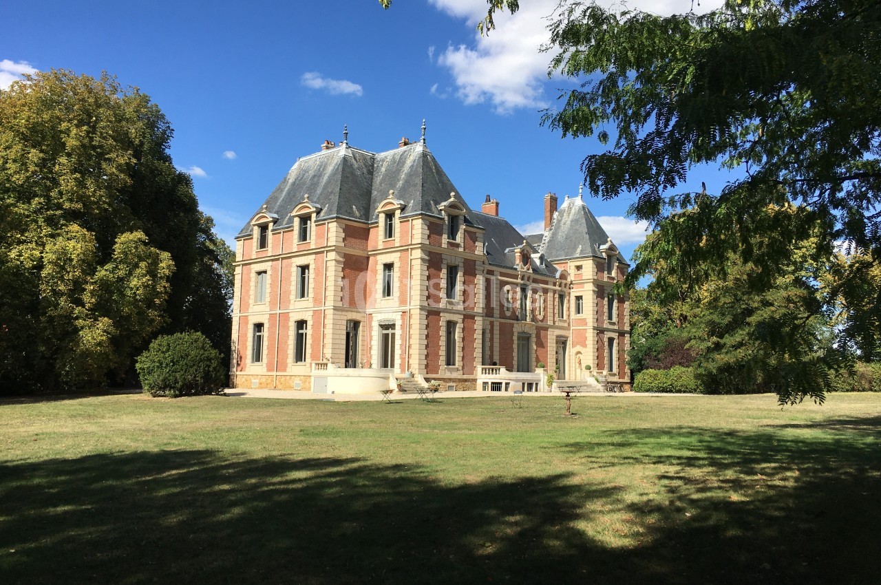 Château en briques et pierres entouré d'un grand parc verdoyant sous un ciel bleu avec quelques nuages.
