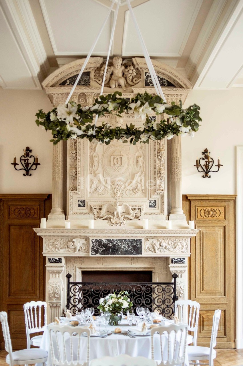 Cheminée sculptée ornée de détails classiques, surmontée d'un lustre floral suspendu, avec table dressée au premier plan.