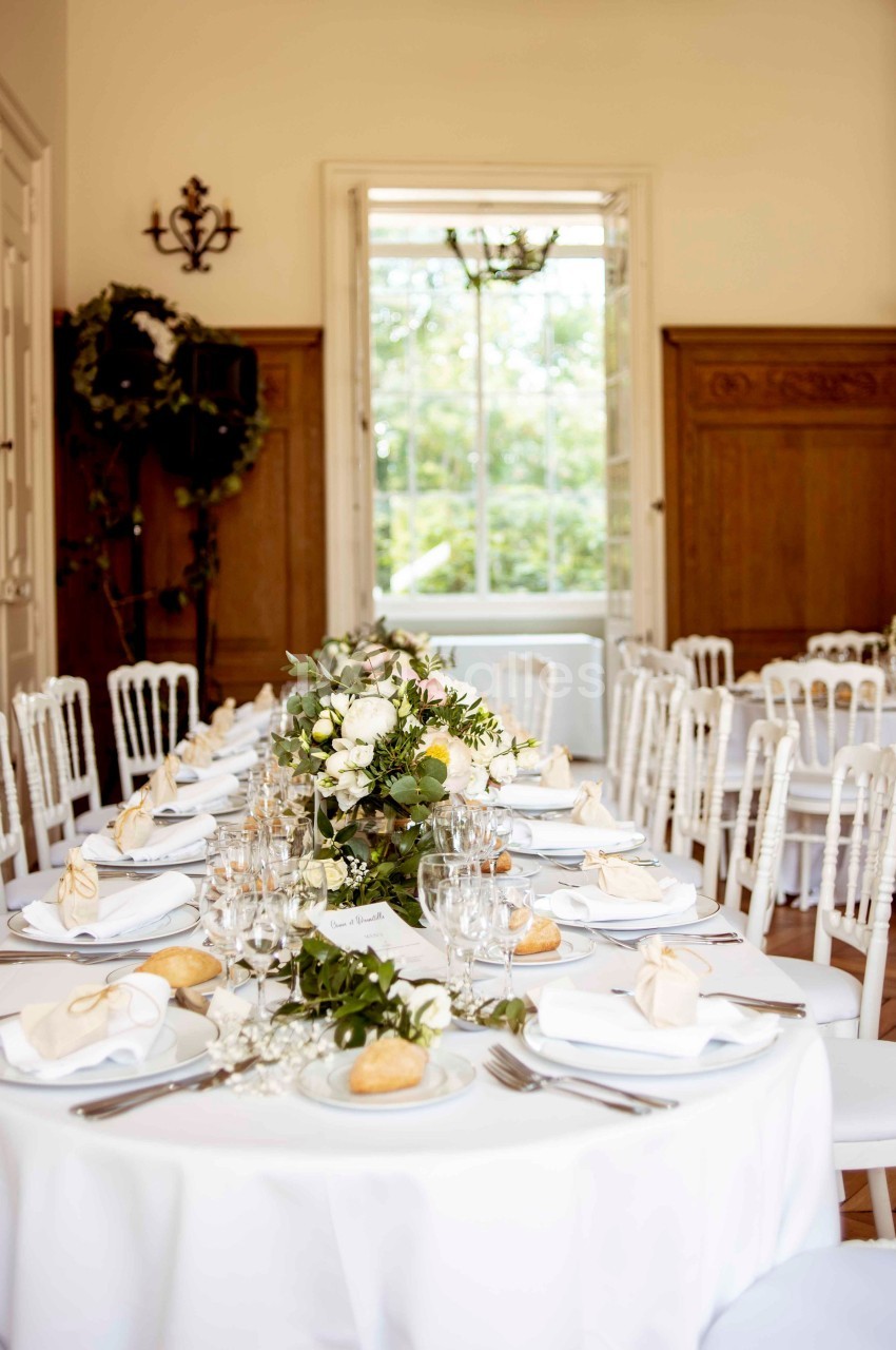 Salle décorée pour un mariage avec des tables rondes dressées, nappes blanches, fleurs et lumière naturelle.