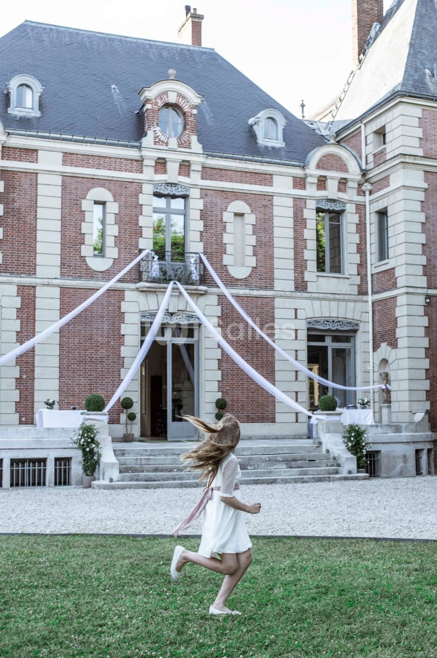 Une fillette en robe blanche court sur une pelouse devant une grande bâtisse en briques décorée pour un événement.