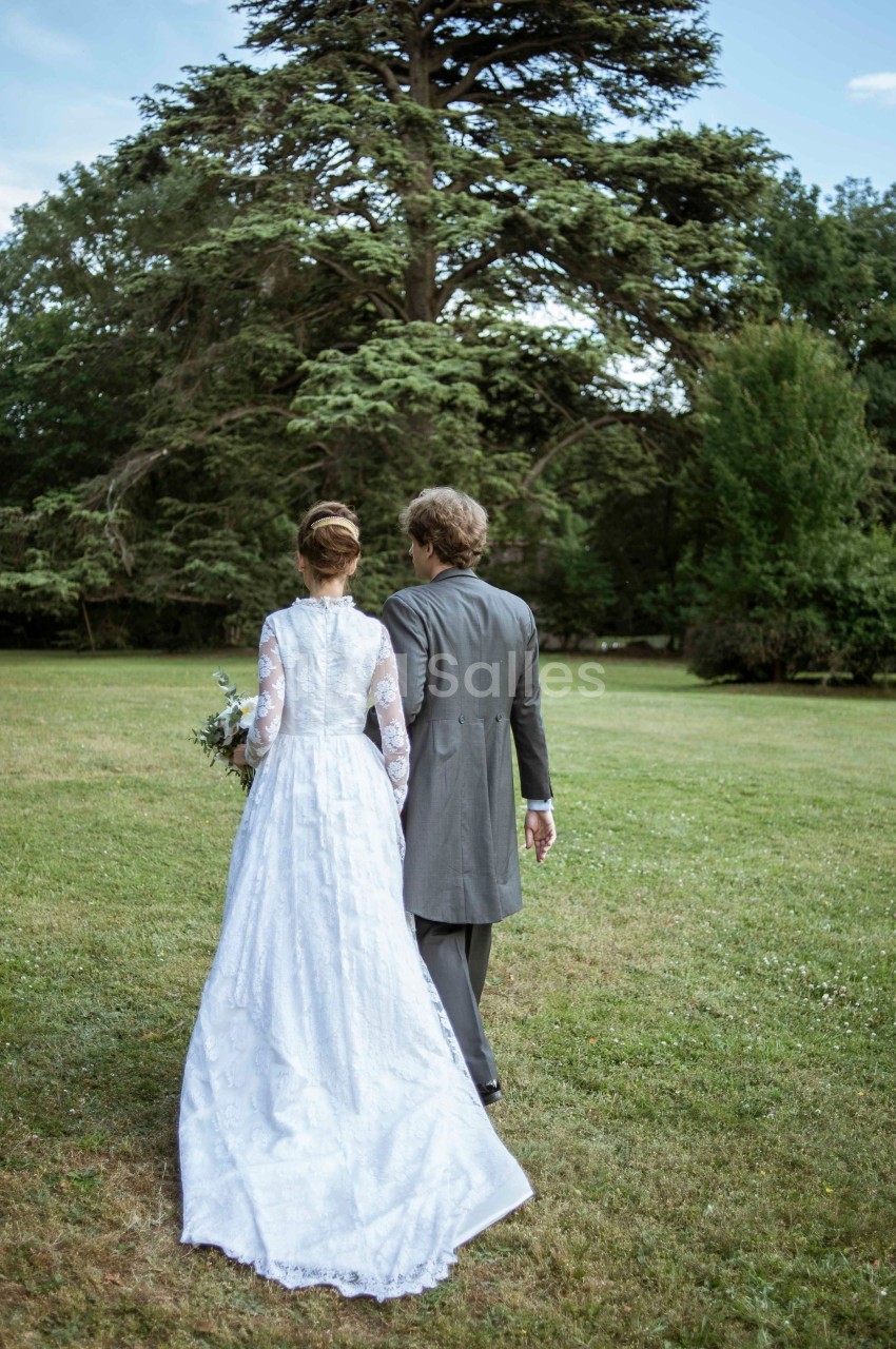 Un couple en tenue de mariage marche dans un parc verdoyant, vu de dos, sous un grand arbre.