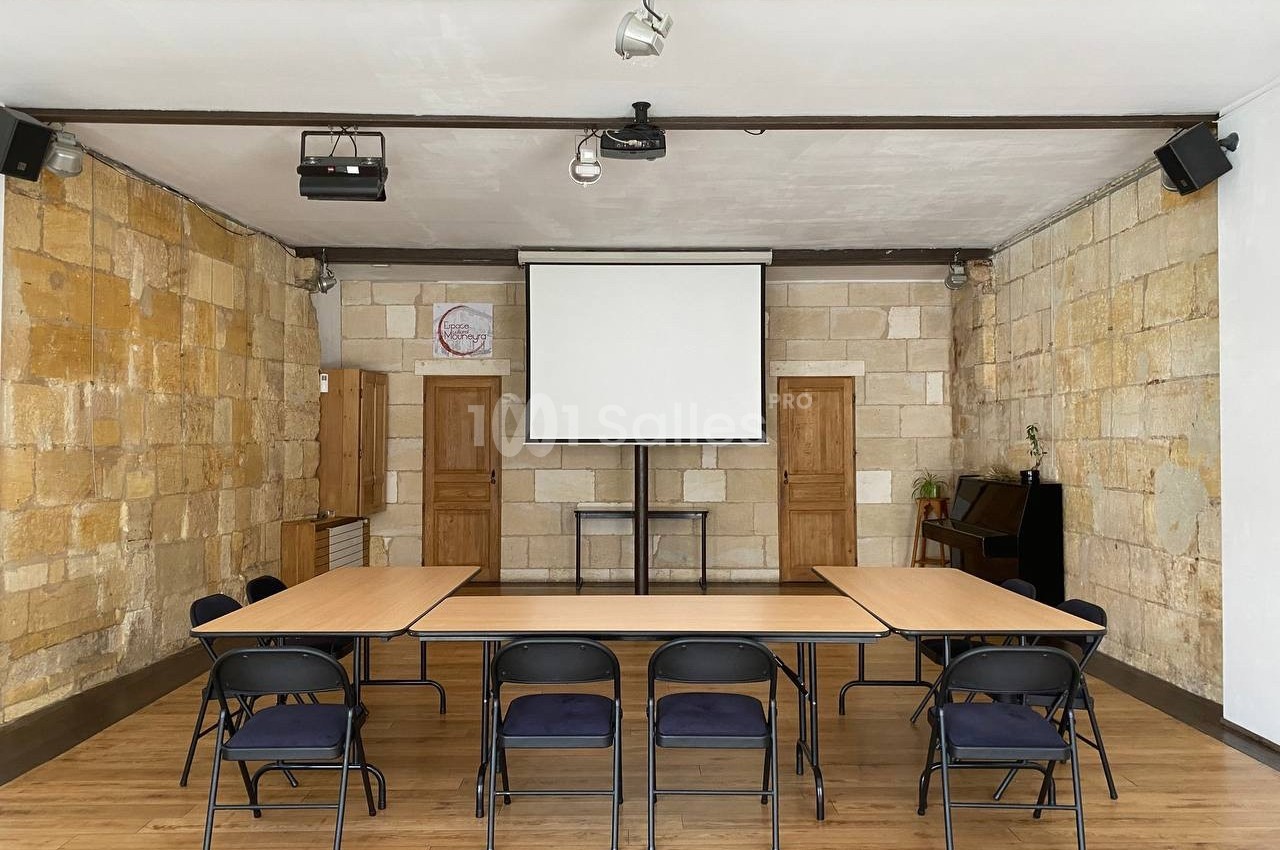 Salle de réunion avec table rectangulaire, chaises pliantes, mur en pierre, écran de projection et parquet en bois.