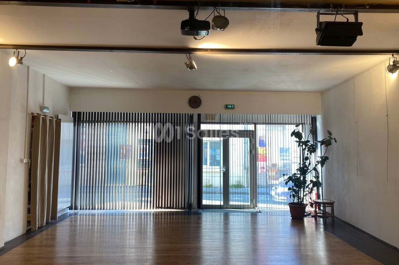 Salle lumineuse avec parquet, grande baie vitrée équipée de stores verticaux, et une plante en pot près de l'entrée.