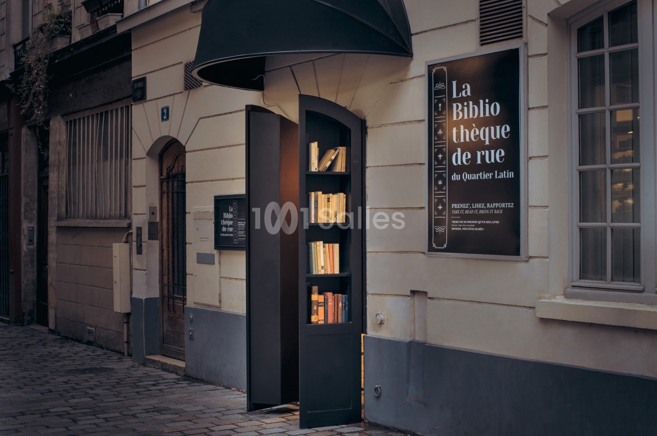 Une petite bibliothèque de rue intégrée dans un mur, éclairée, avec des livres visibles sous un auvent noir.