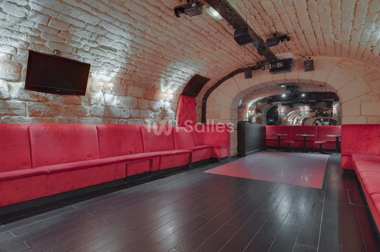 Salle voûtée en pierre avec banquettes rouges, sol en bois sombre et éclairage tamisé.