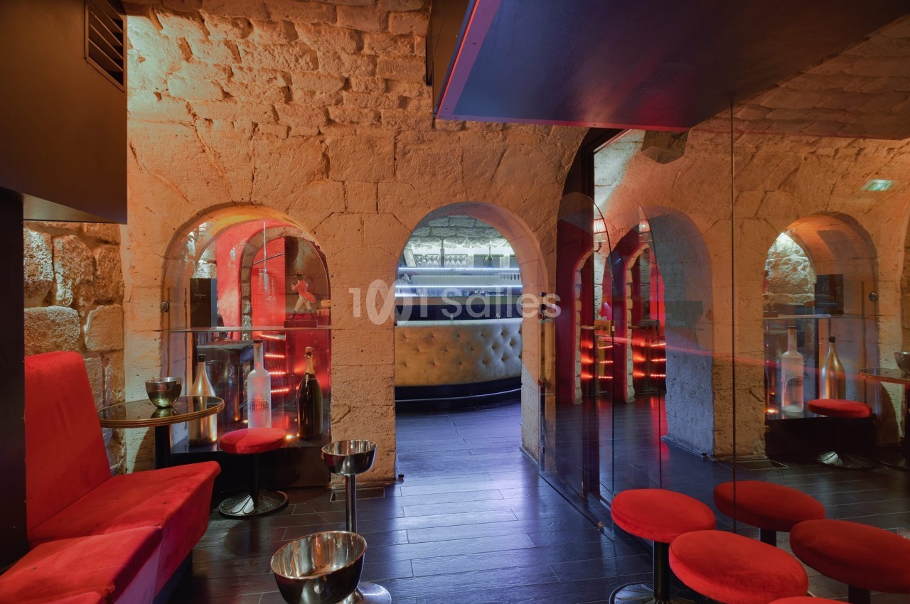 Salle voûtée avec murs en pierre, banquettes rouges, tabourets, et éclairage tamisé près d'un bar moderne.