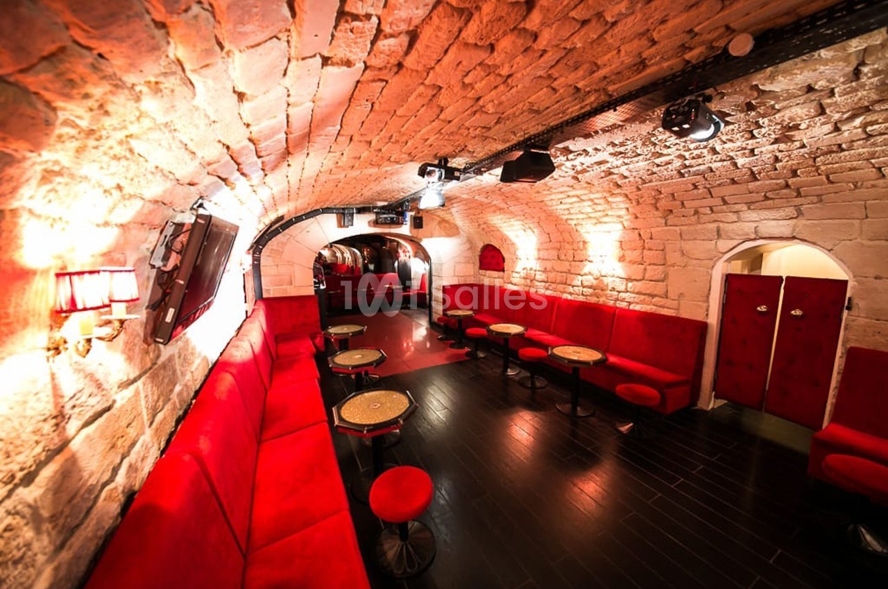 Salle voûtée en pierre avec banquettes rouges, tables rondes et éclairage tamisé.