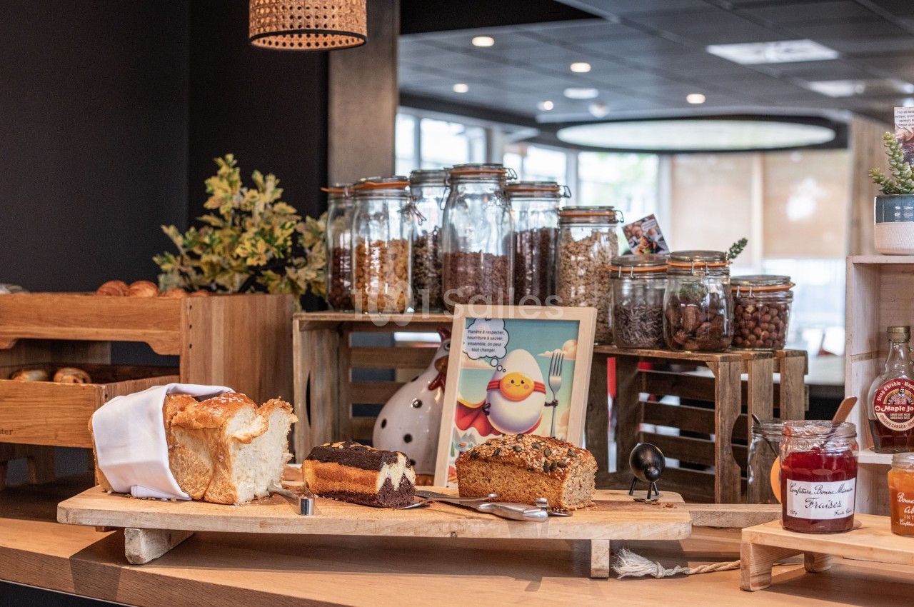 Buffet avec pains, gâteaux, confitures et bocaux de céréales sur une table en bois dans un espace lumineux.