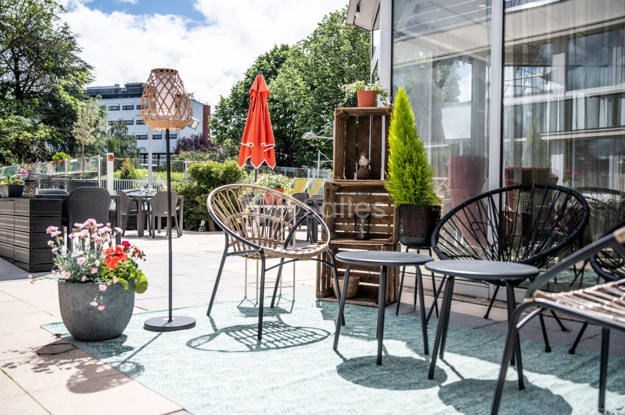 Terrasse aménagée avec des chaises en rotin, une table basse, des plantes en pot et un parasol rouge sous un ciel ensoleillé.