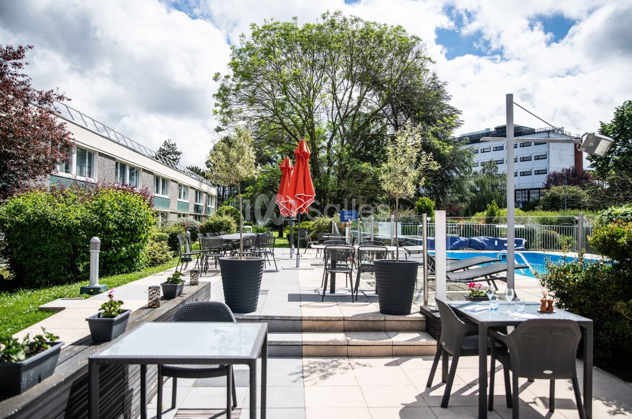 Terrasse extérieure avec tables, chaises, parasol rouge, piscine et espaces verts sous un ciel partiellement nuageux.