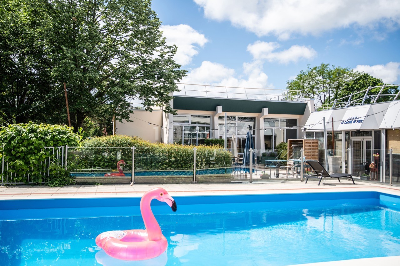 Piscine extérieure avec un flamant rose gonflable, entourée de verdure et d'un bâtiment moderne en arrière-plan.