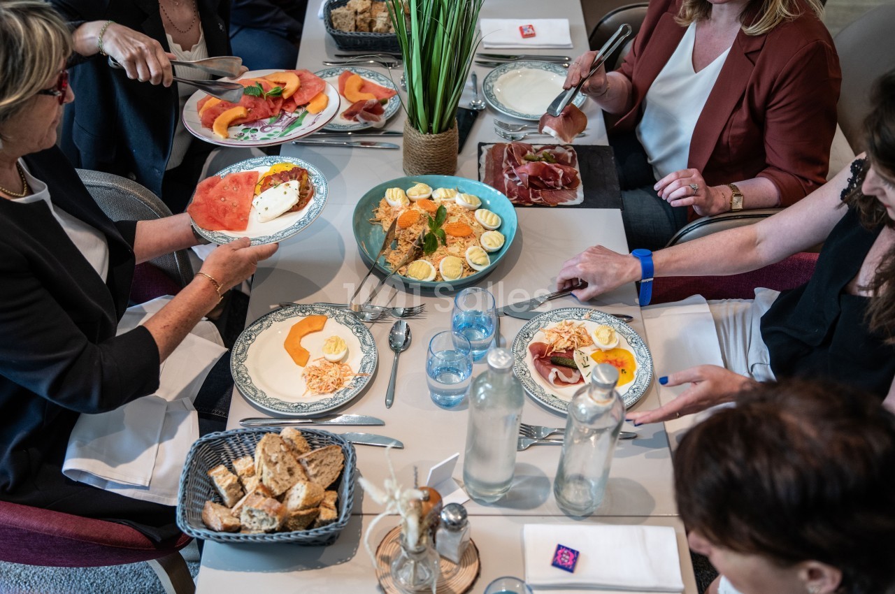 Un groupe de personnes partage un repas composé de fruits, œufs, charcuterie et pain autour d'une table dressée.
