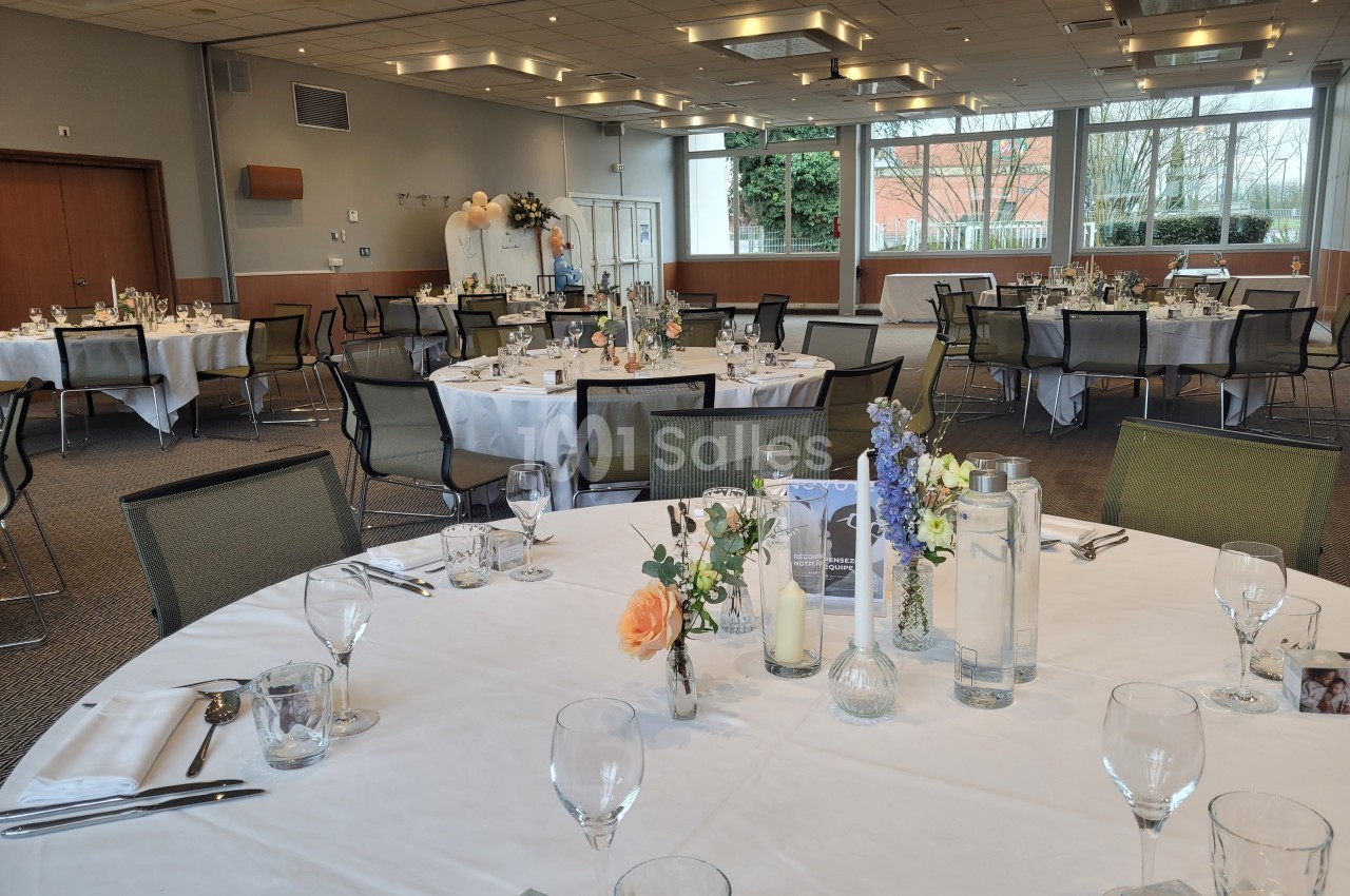 Salle de réception décorée avec des tables rondes dressées, ornées de fleurs et de bougies, prête pour un événement.