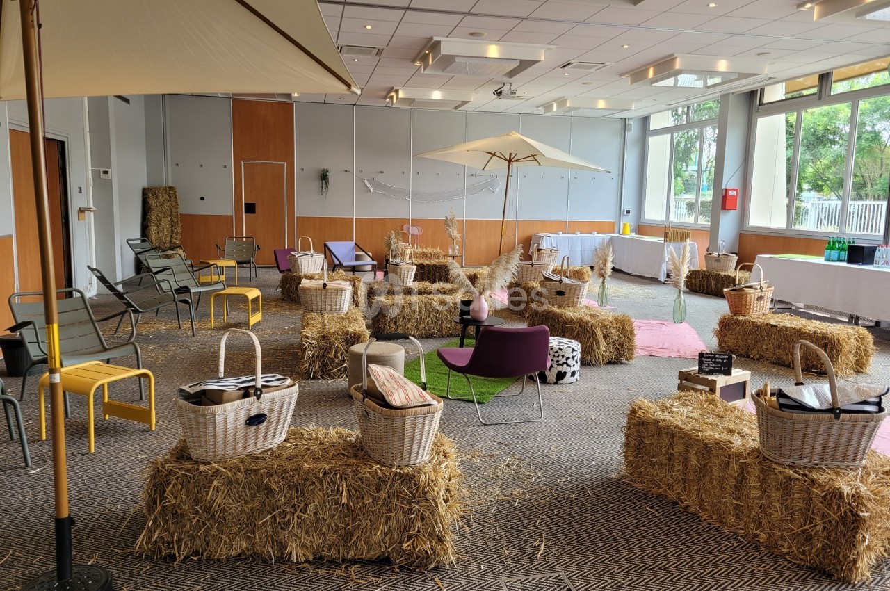 Salle aménagée avec des bottes de paille, des parasols, des chaises et des paniers, dans un cadre lumineux et convivial.