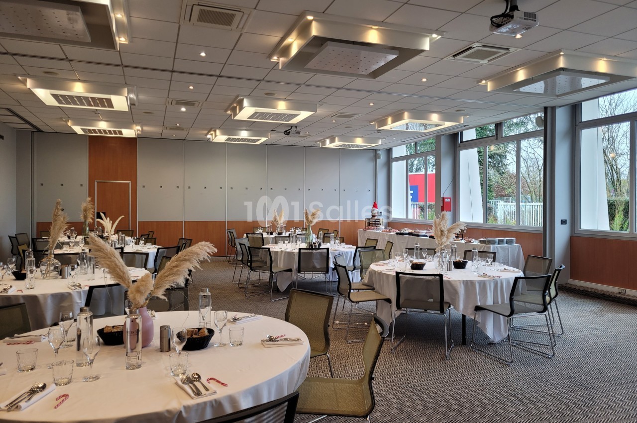 Salle de réception lumineuse avec tables rondes dressées, décorées de plumes blanches, et grandes fenêtres donnant sur l…
