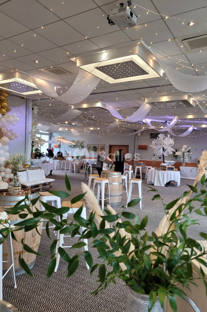 Salle décorée pour un événement avec guirlandes lumineuses, tables dressées et éléments floraux.