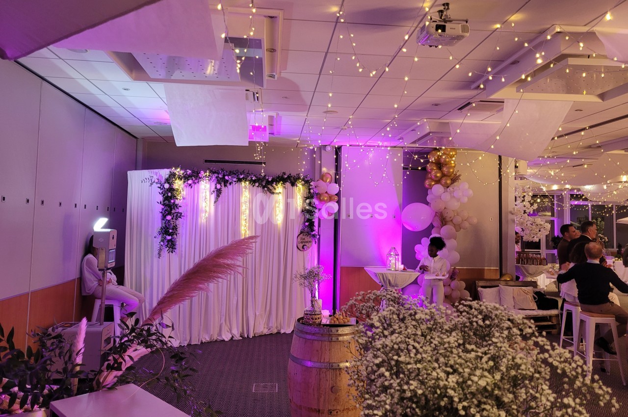 Salle décorée pour un événement avec guirlandes lumineuses, rideaux blancs, fleurs et tonneaux en bois.