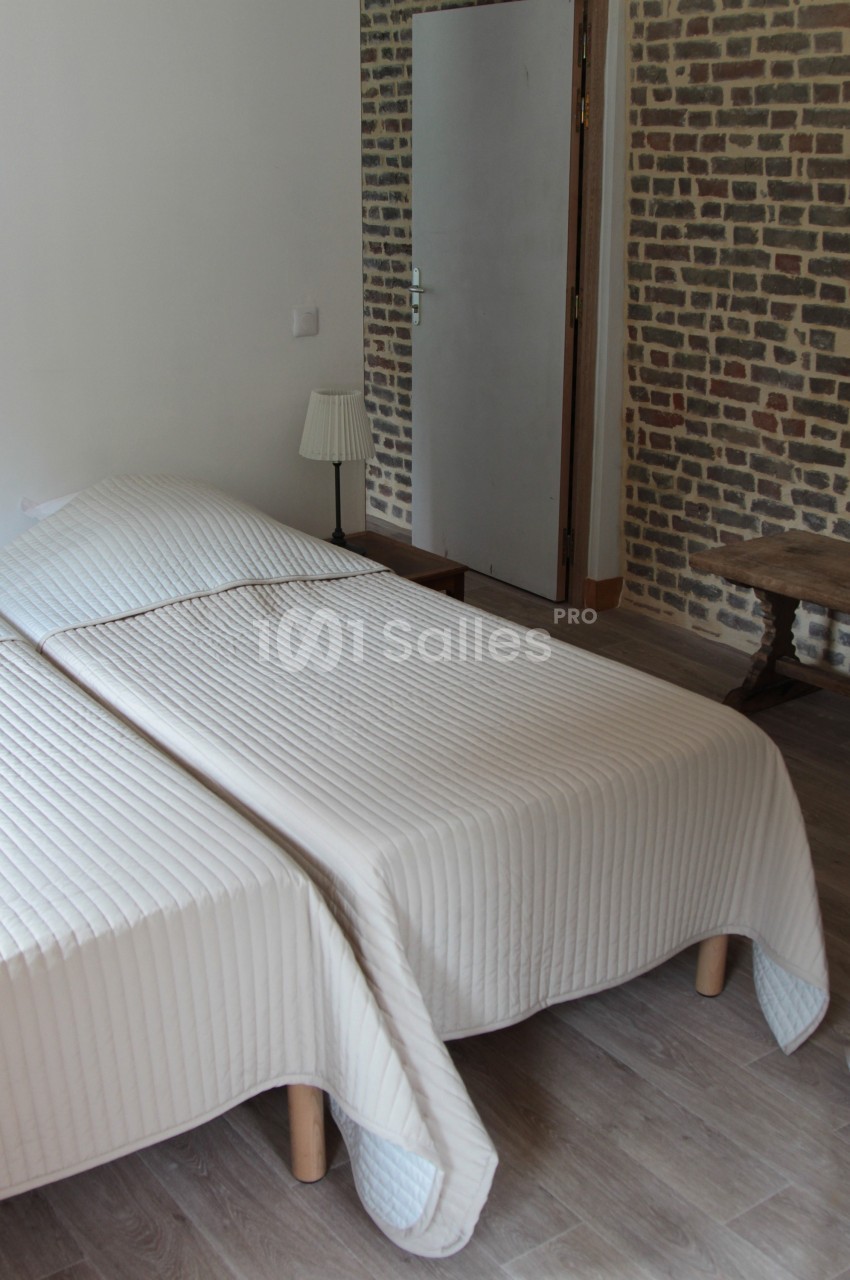 Deux lits simples rapprochés avec des couvre-lits blancs dans une chambre aux murs en briques et sol en parquet.