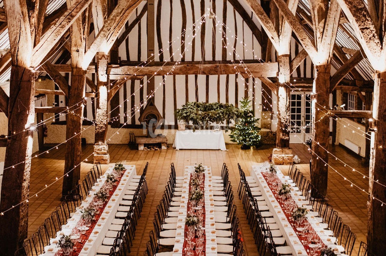 Grande salle de réception avec poutres apparentes, tables longues décorées et guirlandes lumineuses suspendues.