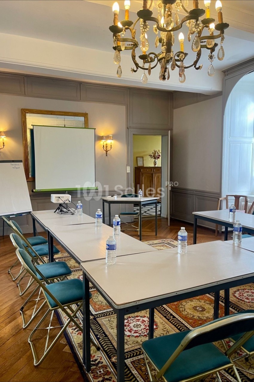Salle de réunion lumineuse avec tables disposées en U, chaises pliantes, tableau blanc, écran de projection et bouteilles d…