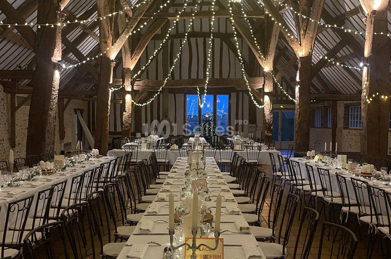 Grande salle de réception rustique avec poutres apparentes, tables dressées et guirlandes lumineuses suspendues.