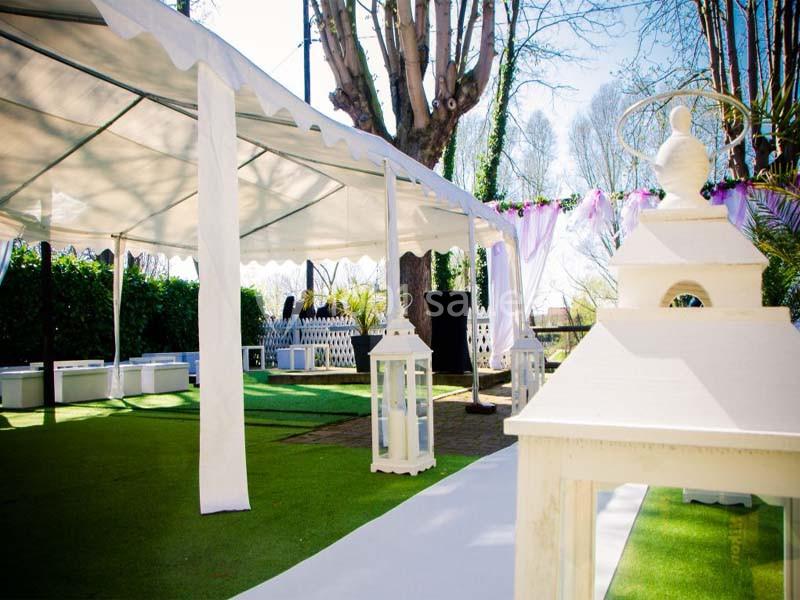 Tente blanche installée dans un jardin avec allée décorée et lanternes blanches, entourée de verdure.