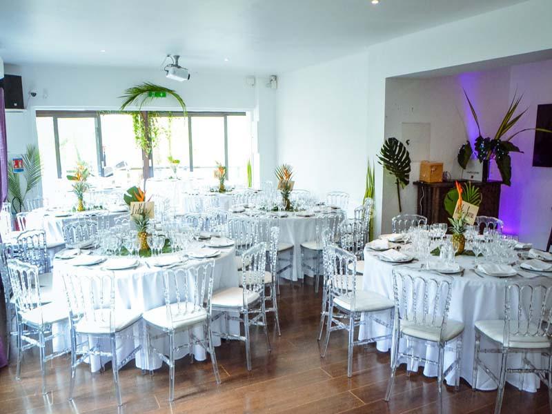 Salle de réception lumineuse avec tables rondes dressées, chaises transparentes et décorations végétales.