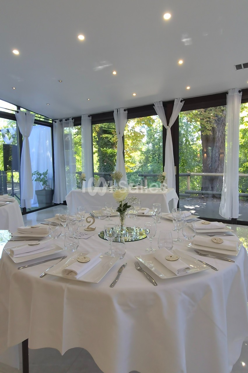 Salle de réception lumineuse avec tables rondes dressées, nappes blanches et vue sur un jardin arboré.