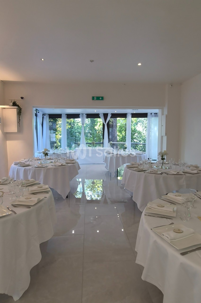 Salle de réception lumineuse avec tables rondes dressées, nappes blanches et vue sur un jardin à travers de grandes baies…