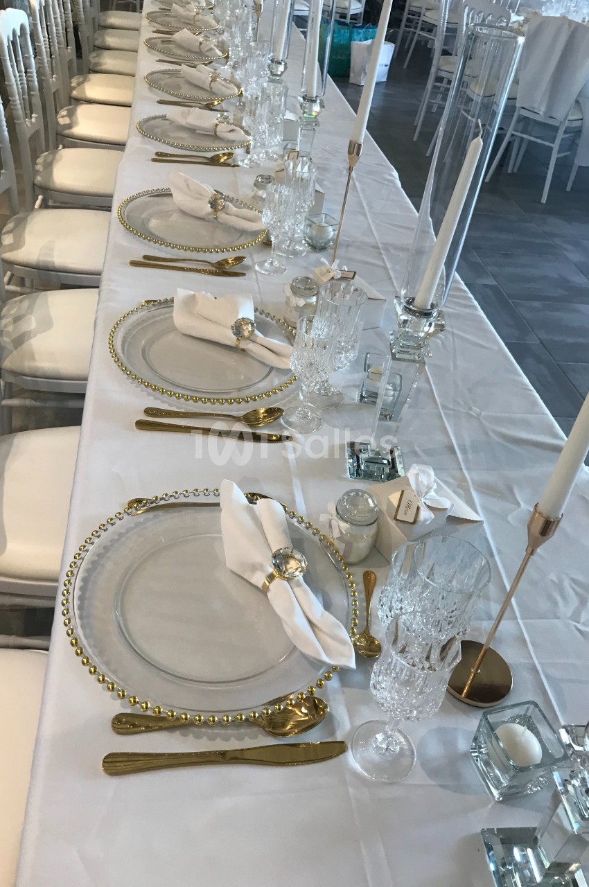 Table élégamment dressée avec assiettes transparentes, couverts dorés, serviettes pliées et chandeliers blancs.