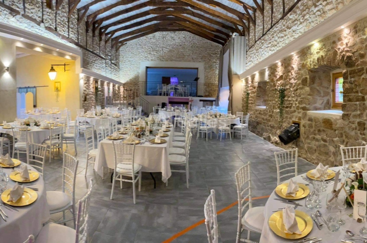 Salle de réception en pierre avec tables rondes dressées, nappes blanches et décoration élégante sous un plafond en poutres…