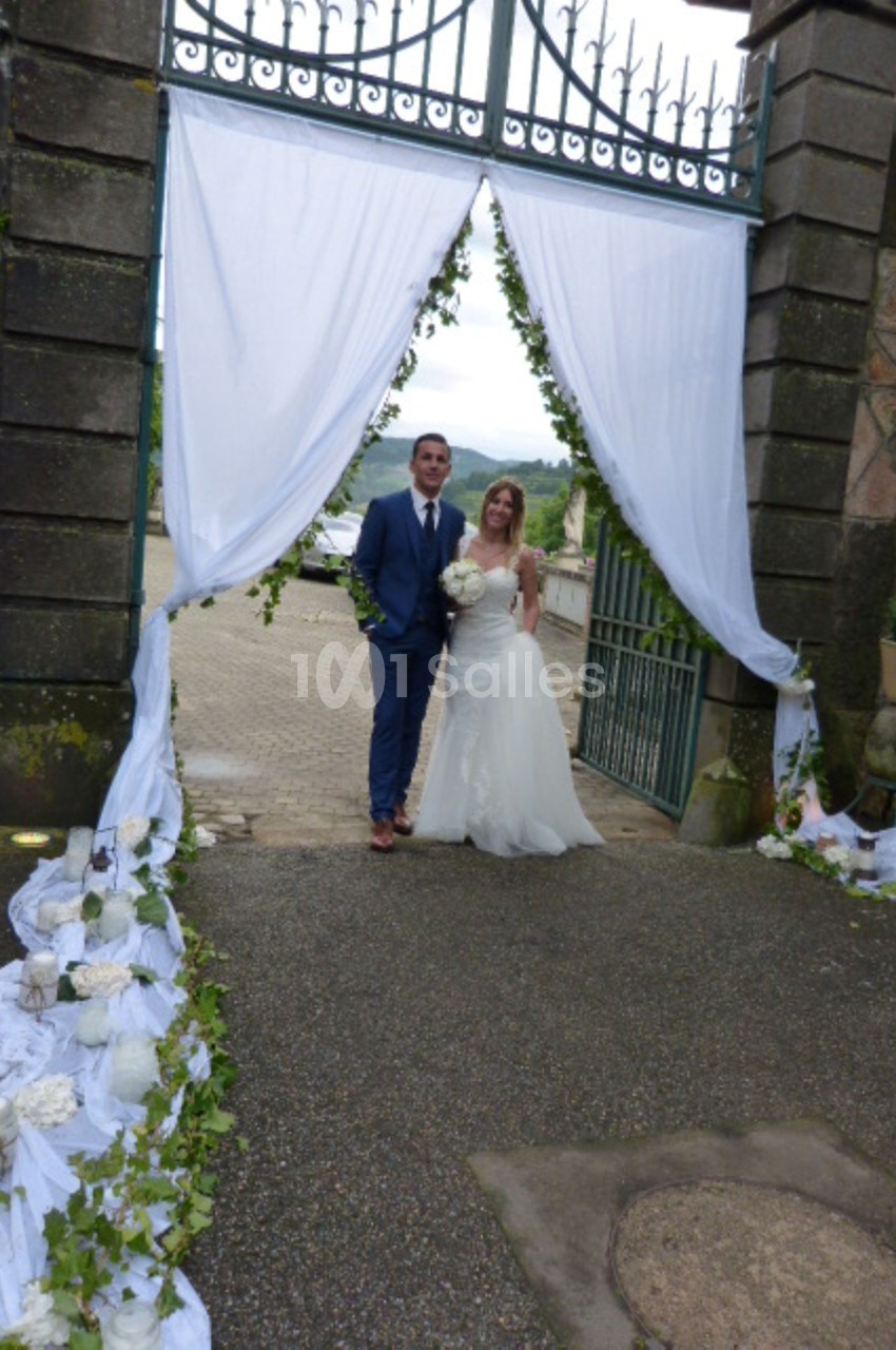 Un couple en tenue de mariage pose sous une arche décorée de drapés blancs et de fleurs, à l'extérieur.