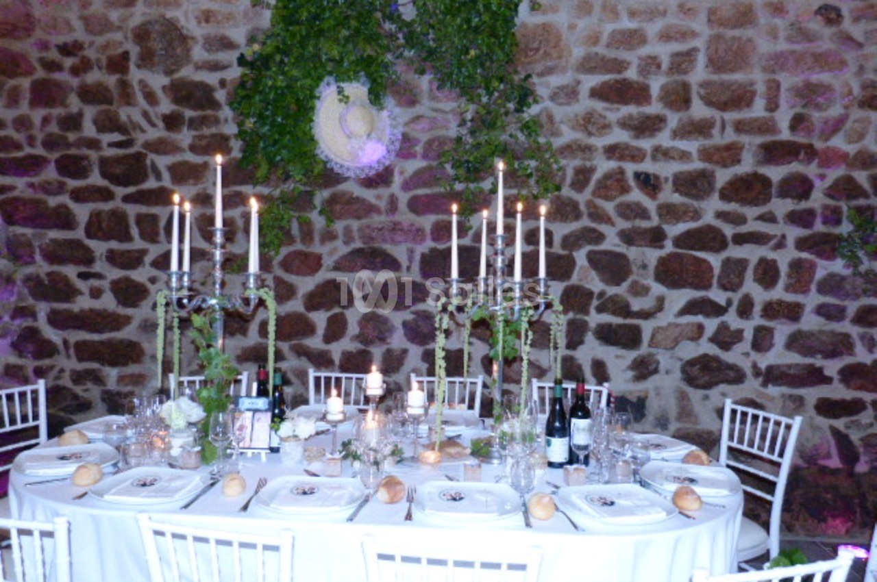 Table élégamment dressée avec chandeliers, vaisselle raffinée et décor végétal devant un mur en pierre.