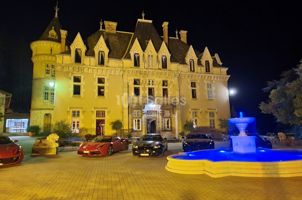 Façade éclairée d'un château de nuit avec une fontaine illuminée au premier plan et des voitures garées devant.