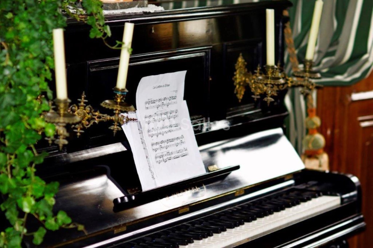 Piano droit noir avec des partitions posées sur le pupitre, entouré de chandeliers et de plantes décoratives.