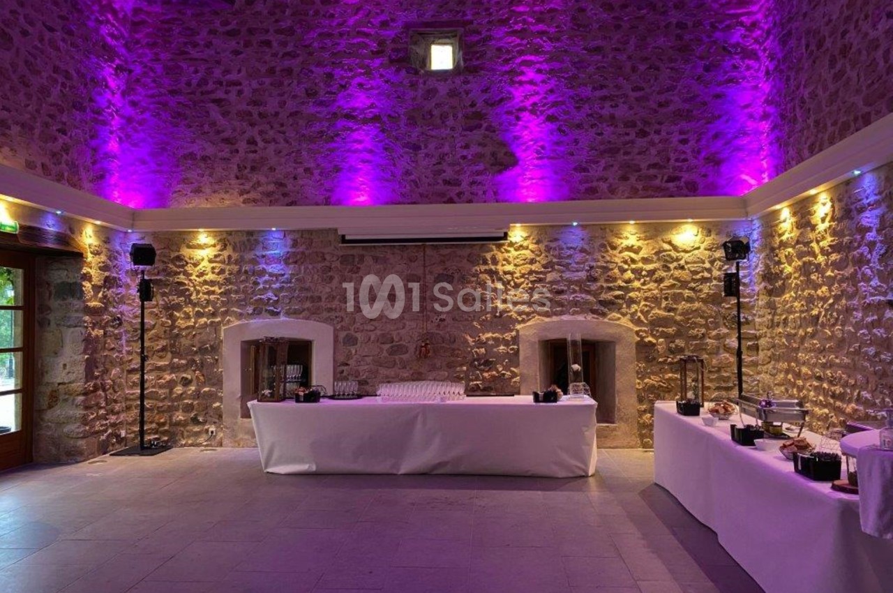 Salle en pierre avec éclairage violet, tables dressées avec nappes blanches et décorations pour un événement.