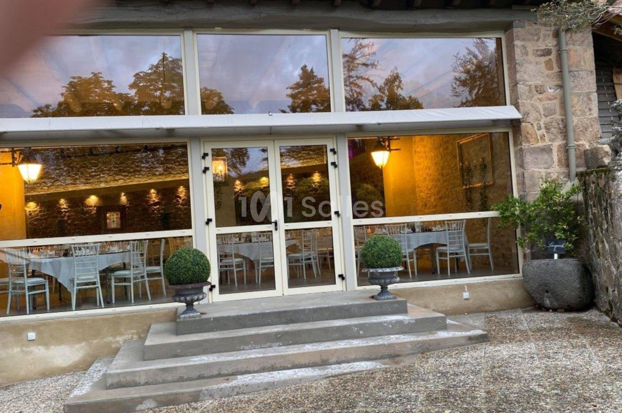 Entrée vitrée d'un bâtiment en pierre avec des tables dressées visibles à l'intérieur et des arbustes décoratifs devant.