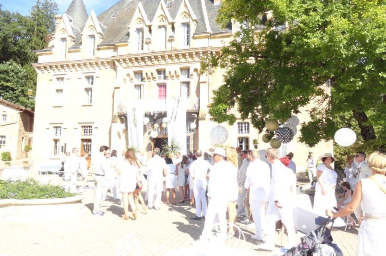 Groupe de personnes vêtues de blanc rassemblées devant un château dans une cour ensoleillée avec des décorations festives.
