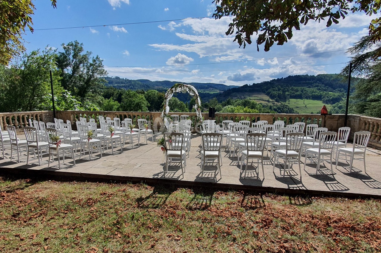 Terrasse panoramique avec chaises blanches, arche florale et vue sur la vallée verdoyante des montagnes.