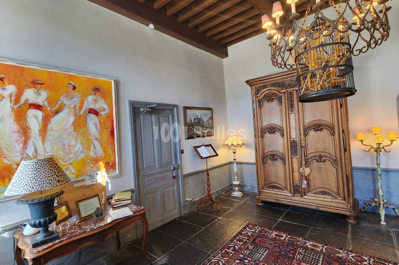 Entrée du château avec plafond à poutres apparentes, tableau moderne aux tons orange, mobilier ancien et cage dorée suspendue.