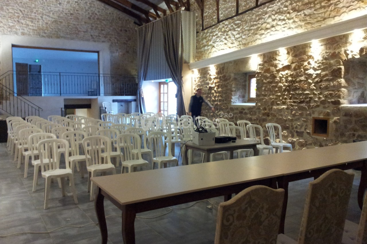 Salle en pierre avec plafond en poutres apparentes, rangées de chaises blanches et tables installées pour un événement.