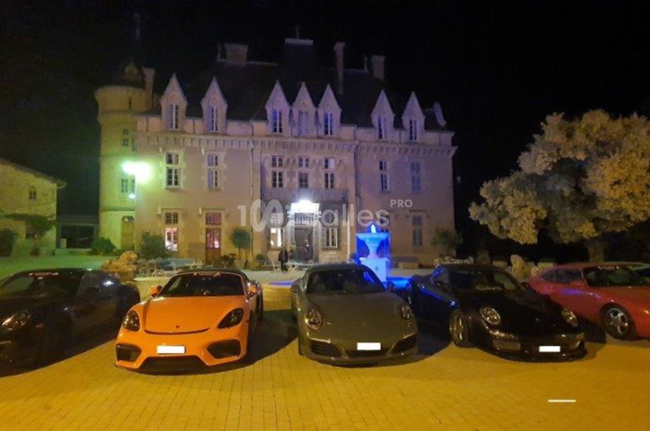 Cinq voitures de sport alignées devant un château éclairé la nuit, avec une fontaine illuminée au centre.