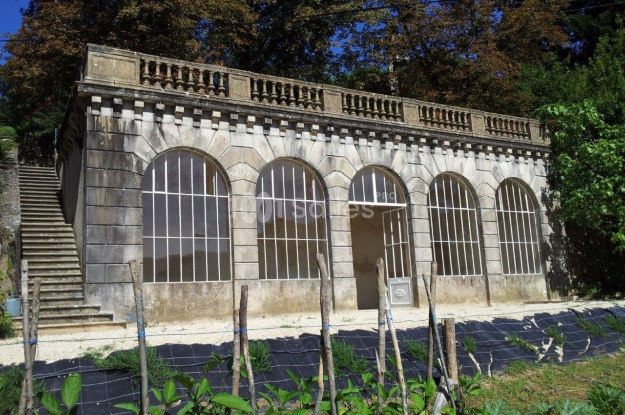 Façade d'un bâtiment ancien avec des arches vitrées, entouré de végétation et d'un escalier sur la gauche.