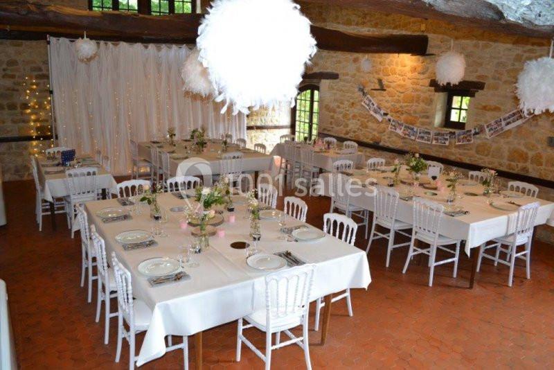 Salle de réception décorée avec des tables dressées, des chaises blanches et des guirlandes lumineuses sur un mur en pierre.