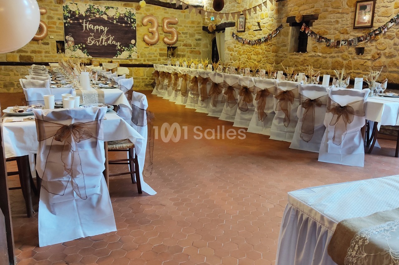 Salle décorée pour un anniversaire avec des tables alignées, nappes blanches et chaises ornées de nœuds marron.