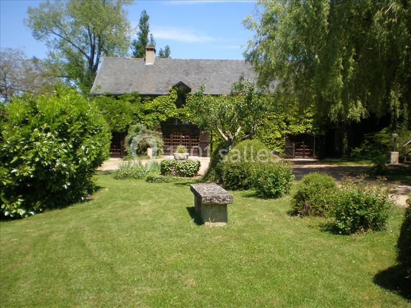 Maison en pierre couverte de lierre, entourée d'un jardin verdoyant avec pelouse, arbres et banc en pierre.