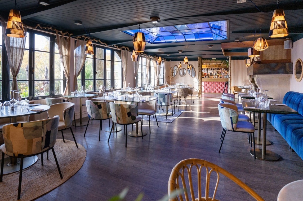 Salle de restaurant lumineuse avec grandes baies vitrées, mobilier moderne et bar en arrière-plan.