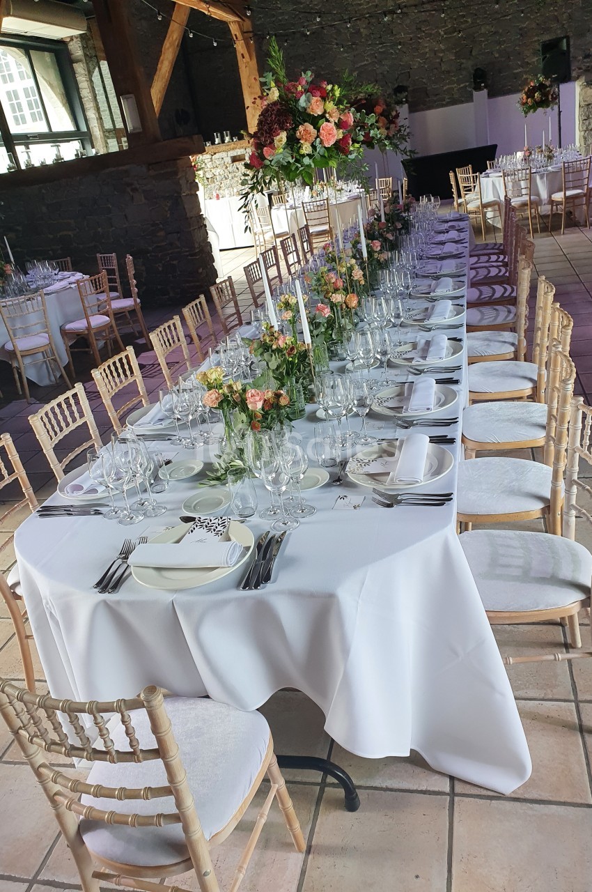 Table dressée pour un événement, avec nappes blanches, chaises en bois, vaisselle élégante et décorations florales colorées.