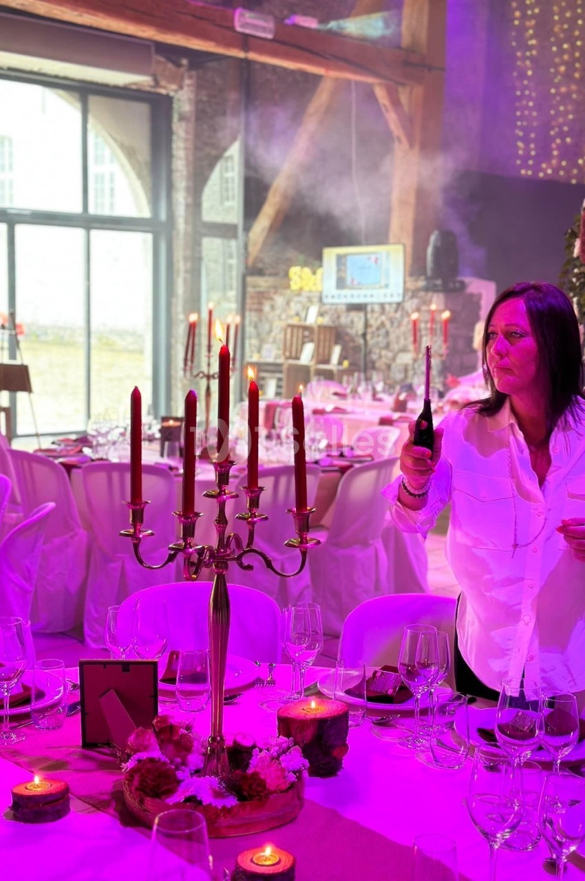 Une femme allume un chandelier à bougies dans une salle décorée pour un événement, avec des tables dressées.