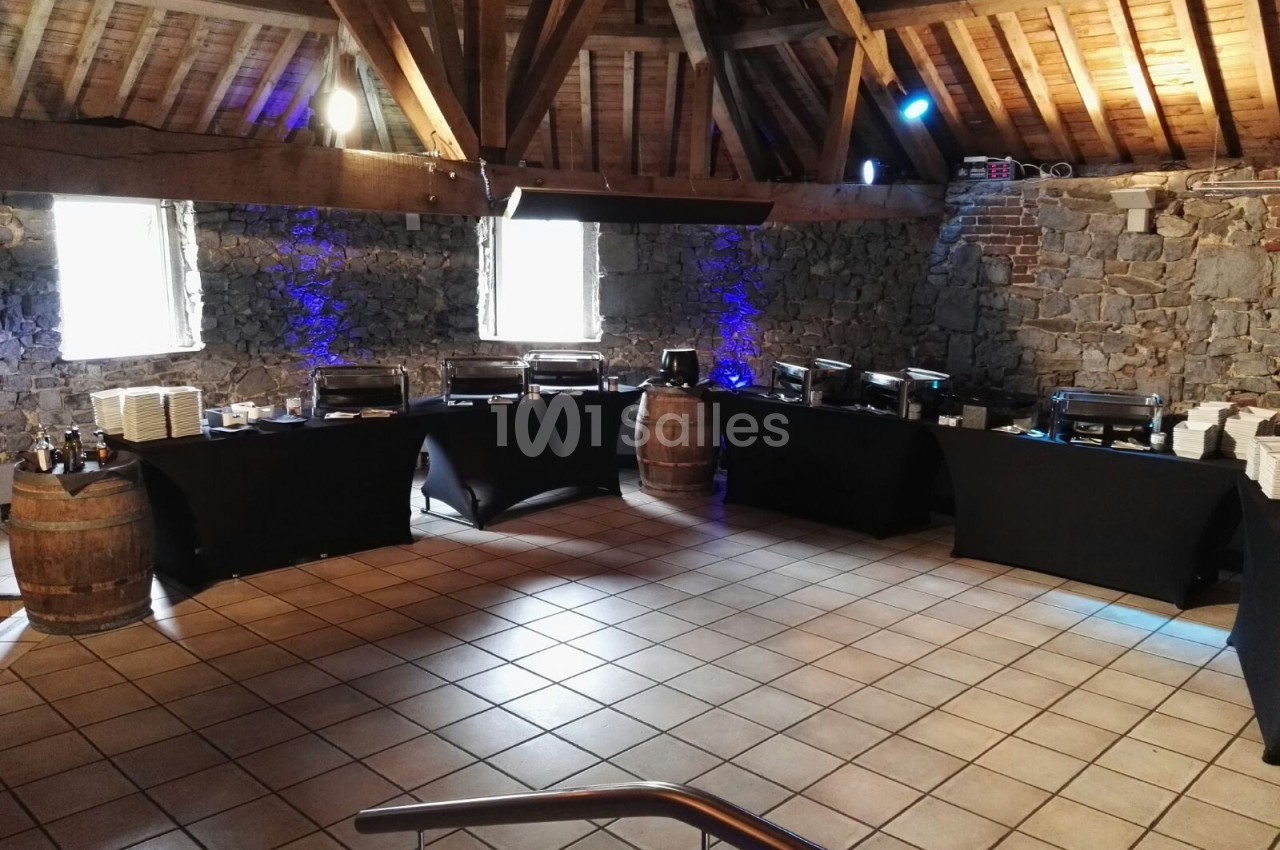 Salle en pierre avec plafond en poutres apparentes, tables noires alignées pour un buffet, éclairage tamisé.